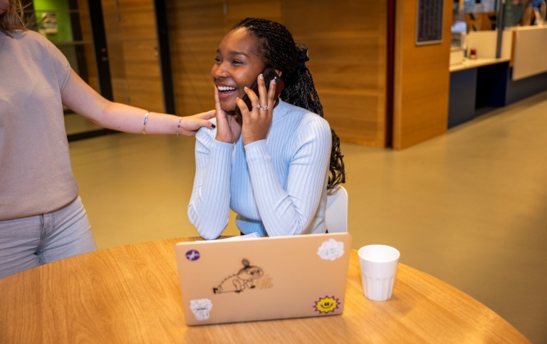 Glad kvinna som pratar i telefon. En kvinna står bredvid och håller en hand på hennes axel.